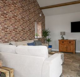 A living room with a beige sofa set against a brick wall a wooden cabinet with a lamp and a mounted black TV at Owl View in Lebberston near Cayton