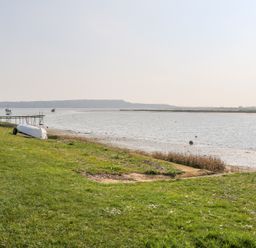 A grassy shoreline with a small boat on the shore a wooden pier and boats in the water at Harbour View Mudeford in Christchurch