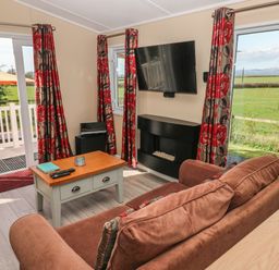 A living room with a sofa coffee table wall-mounted TV and windows overlooking a garden at Mountain View Lodge Tavernspite near Narberth