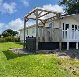 A yard with a wooden pergola and hot tub next to a cream-colored house with white railings at Mountain View Lodge Tavernspite near Narberth