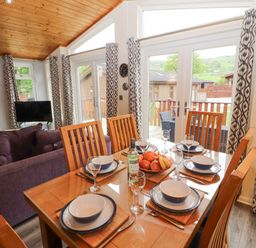 A dining table set with plates and glasses next to a living area with purple sofas and large windows with patterned curtains at Fell View Lodge in Troutbeck