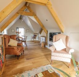 A living area with wooden beams a brown armchair a pink patterned armchair a bookshelf and a dining table at The Coach House in St Columb Major