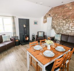 A living room with two brown leather sofas a wooden dining table set with plates and glasses a stone wall and a wood burning stove at Dairy Cottage Tavernspite near Narberth