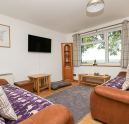 A living room with two brown sofas purple patterned throws wooden tables a wall mounted TV and a window with plaid curtains at Dartmoor 2 in Callington