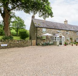 A stone cottage with outdoor seating and potted plants beside a gravel parking area at Isaacs Cottage Spartylea near Allendale