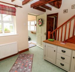 An entrance hallway with a wooden staircase a cabinet with drawers and flowers on top a window with a floral vase and a doorway leading to another room at Isaacs Cottage Spartylea near Allendale