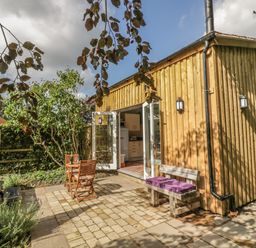 A wooden exterior building with open glass doors leading to a kitchen and a patio area with a wooden table and chairs and a bench with purple cushions at Hen House View in Rugeley
