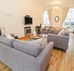 A living room with grey sofas a round wooden coffee table a wall-mounted television and large arched windows at Seion Chapel in Beaumaris