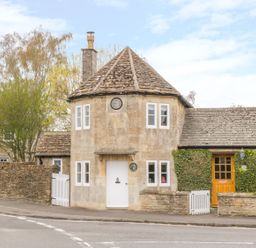 A stone house with a conical roof a white door and clock on the wall surrounded by a stone wall and greenery at Pike Cottage in Acton Turville near Chipping Sodbury
