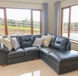 A corner black leather sofa with cushions in a room with large windows and grey curtains at Rossanean in Farranfore near Killarney County Kerry