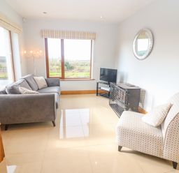 A sitting room with a gray sofa, patterned chair, TV on stand, and large windows at Rossanean in Farranfore near Killarney County Kerry