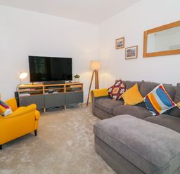 A living room with a grey sofa and yellow armchair facing a TV on a wooden stand at Lyngaran House in Holyhead