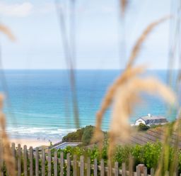 A view of the ocean with waves and a sandy beach behind a wooden fence and grass at Blue Bay Beach House in Mawgan Porth