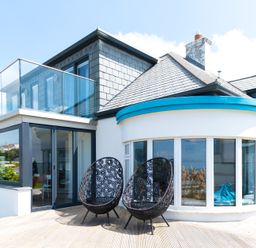 An outdoor patio with two black decorative chairs next to a modern house with a glass balcony and large windows at Blue Bay Beach House in Mawgan Porth