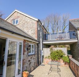 A stone building with a glass door opening to a patio with a small table and a balcony with outdoor furniture at Lambkin Cottage in St Columb Major
