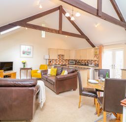 An open plan living room with brown leather sofas a yellow chair a wooden dining table with four chairs a kitchen area with light wood cabinets and tiled backsplash at Lambkin Cottage in St Columb Major