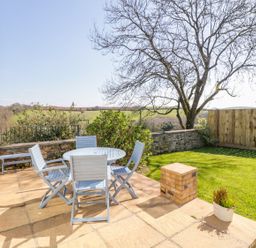 An outdoor patio with a round table and four chairs next to a lawn with a tree and a wooden fence at Forget-me-not in St Columb Major