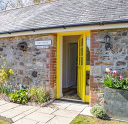 An entrance with a yellow door in a stone building with flowers in a planter and garden bed at FernGully in St Columb Major