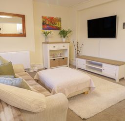 A living room with a sofa and ottoman a TV mounted on the wall and a cabinet with plants and decorations at Beside The Sea - In Town in Llandudno
