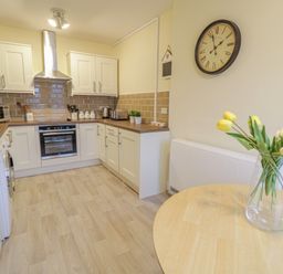 A kitchen with white cabinets brown tiled backsplash and a wooden table with yellow flowers in a vase at Beside The Sea - In Town in Llandudno