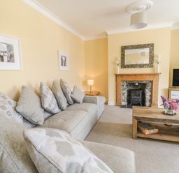 A living room with a grey sofa and multiple cushions a wooden coffee table with flowers a stone fireplace and a television on a stand at Angorfa in Llanbedr
