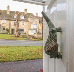 A fish shaped door knocker on a white door with stone buildings and grass across the road at Dolphin House in Burford