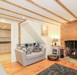 A living room with a sofa, pillows, a wood burning stove in a brick fireplace, a small table with a lamp and books and a view into a kitchen at Tilly's Cottage in Edington