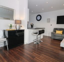A living area with a gray sofa, yellow pillows, a wall-mounted television, two black bar stools at a counter, a black cabinet with a table lamp and flowers, and white chairs at 8 Marina View in Pwllheli