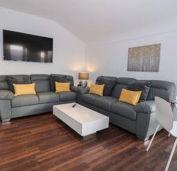 A living room with gray sofas yellow and black cushions a white coffee table a wall-mounted television a lamp and a glass dining table with white chairs at 8 Marina View in Pwllheli