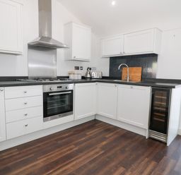 A kitchen with white cabinets black countertops a stainless steel oven and stove a sink a cutting board a kettle a toaster a wine fridge and wood flooring at 8 Marina View Pwllheli
