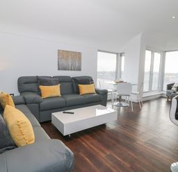 A living room with grey sofas and yellow cushions a white coffee table dark wooden floor and a dining table with white chairs at 8 Marina View in Pwllheli