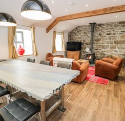 A living room with a stone wall fireplace brown armchairs a dining table with black chairs and two pendant lights at Rhydau Cottage in Pentir near Deiniolen