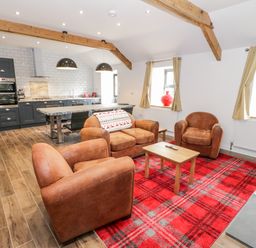 A seating area with brown armchairs and a sofa on a red plaid rug next to a kitchen with dark cabinets and two pendant lights at Rhydau Cottage in Pentir near Deiniolen