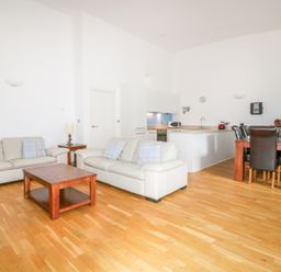 A living room with two cream sofas and a wooden coffee table next to a kitchen with white cabinets and a dining table with six dark chairs at 28 Zenith in Porth