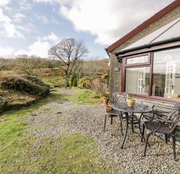 An outdoor seating area with a metal table and two chairs next to a building and a gravel pathway with trees and grass at Melin Newydd in Porthmadog