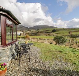 An outdoor seating area with a small table and two chairs next to a stone wall window overlooking grassy fields and hills at Melin Newydd in Porthmadog
