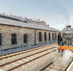 A black steam locomotive on railway tracks next to a stone building with arched windows at Premier, Transit & Lady Hilda @ Engine Shed in Whitby