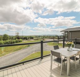 An outdoor deck with a table and four chairs overlooking a gravel road and green landscape at Ash Lodge in Teigngrace near Newton Abbot