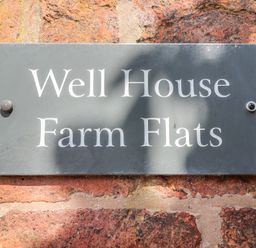 A black sign with white text reading Well House Farm Flats mounted on a red brick wall at Well House Farm Flat 1 in Bretton