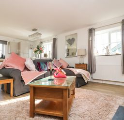 A living room with a gray sofa and pink cushions and throws a wooden coffee table with glass top a dining table with flowers and kitchen area in the background at Be Our Guest Cottage in Chacewater near Truro
