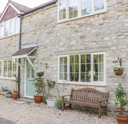 The exterior of a stone house with windows a green door a wooden bench and various potted plants at Duck Cottage in Sutton Poyntz