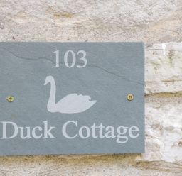 A stone wall with a gray house number plaque showing 103 and a swan above the text Duck Cottage in Sutton Poyntz