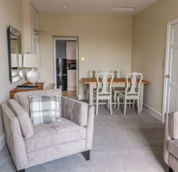 A living room with beige armchairs a wooden dining table with white chairs and a small side table with a lamp at Coquet View Apartment in Warkworth