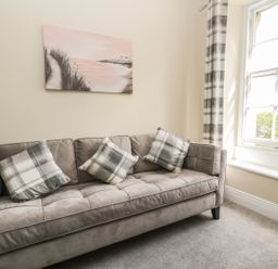 A room with a gray sofa with plaid cushions a window with plaid curtains and a landscape painting on the wall at Coquet View Apartment in Warkworth