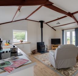 An open plan living area with a kitchen counter a wood stove two armchairs a television and a window with curtains at Village View in Burton Bradstock