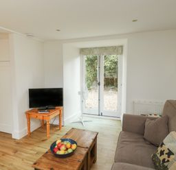 A living room with a brown sofa a wooden coffee table with a bowl of fruit a small wooden table with a television and a standing fan near glass doors at Bantam Cottage in Little Woodford near Ottery St Mary