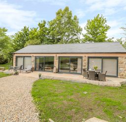 A single story stone house with large sliding glass doors and outdoor seating on a patio surrounded by trees at Coal Drops in Alnwick
