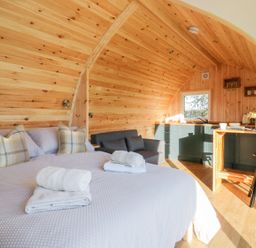 A wooden pod interior with a bed with pillows and towels a small sofa a kitchenette and a dining counter with stools at Chapel Pod in Callington