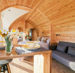 An interior wooden pod with a table set with bowls and plates a gray sofa a bed with pillows and folded towels and a vase of flowers at Chapel Pod in Callington