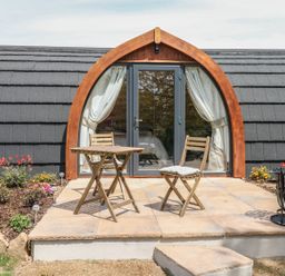 An outdoor patio with a wooden table and two chairs in front of a small cabin with arched glass doors at Chapel Pod in Callington